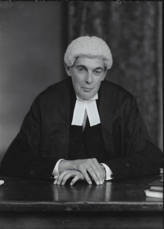 A black-and-white studio portrait of a white British judge from the mid-20th century. He is shown from the waist up, seated at a dark wooden table with hands gently clasped in front. He is wearing traditional legal attire, including a dark robe, white jabot collar bands, and a light-colored judicial wig. He looks directly toward the camera with a faint smile against a dark, softly lit background.