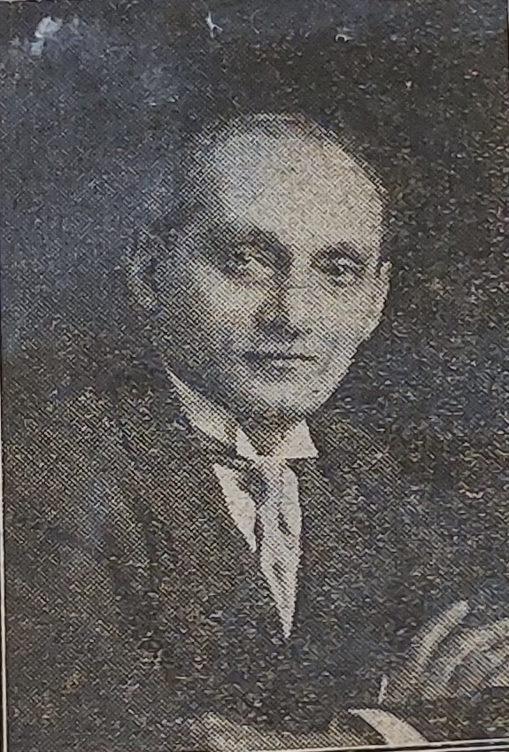 A historical black-and-white studio portrait of an Indian man with dark hair, a high forehead, and a slight smile. He is dressed in formal early-20th-century clothing, including a dark jacket, a white stiff collar shirt, and a wide silk or satin tie. The picture has a coarse, half-tone printing texture typical of old newspaper archival clippings.