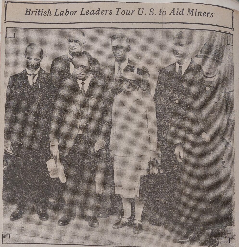 A grainy historical newspaper clipping featuring a group portrait of four men and two women standing outdoors. The men wear formal dark suits and ties, with one holding a light-colored hat. The women wear mid-length coats and cloche hats typical of late 1920s fashion. A headline printed across the top of the photo frame reads: 