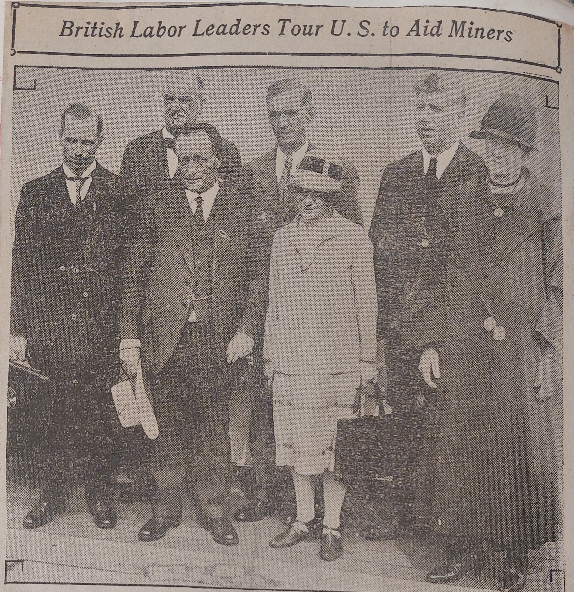 A grainy historical newspaper clipping featuring a group portrait of four men and two women standing outdoors. The men wear formal dark suits and ties, with one holding a light-colored hat. The women wear mid-length coats and cloche hats typical of late 1920s fashion. A headline printed across the top of the photo frame reads: 