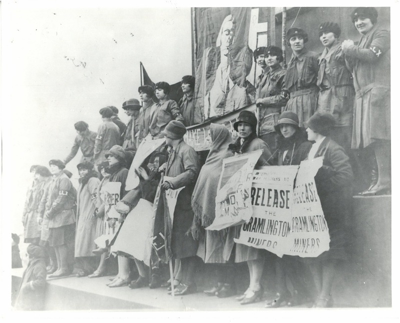 A black and white historical photograph depicting a group of activist women standing on an outdoor elevated platform during a political campaign. Several women in the foreground hold large, hand-painted protest signs that read 