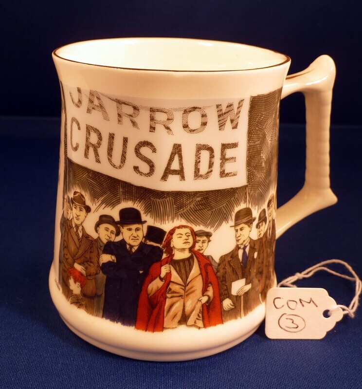 A photograph of a white ceramic commemorative mug featuring an illustration of the 1936 Jarrow Crusade. The illustration wraps around the front of the mug, showing a group of marching figures beneath a large banner that reads 