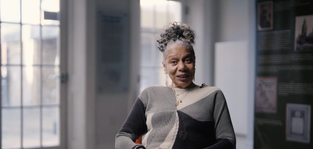 A medium shot of an older Black woman sitting in a chair, appearing to speak while smiling during an interview. She has her hair styled in a high, curly updo with grey streaks. She is wearing a long-sleeved, color-blocked sweater in shades of grey, black, and olive green, layered gold necklaces, and earrings. Her hands are clasped in her lap. The background is a softly blurred indoor setting with a large glass-paned door on the left and an exhibition banner on the right.