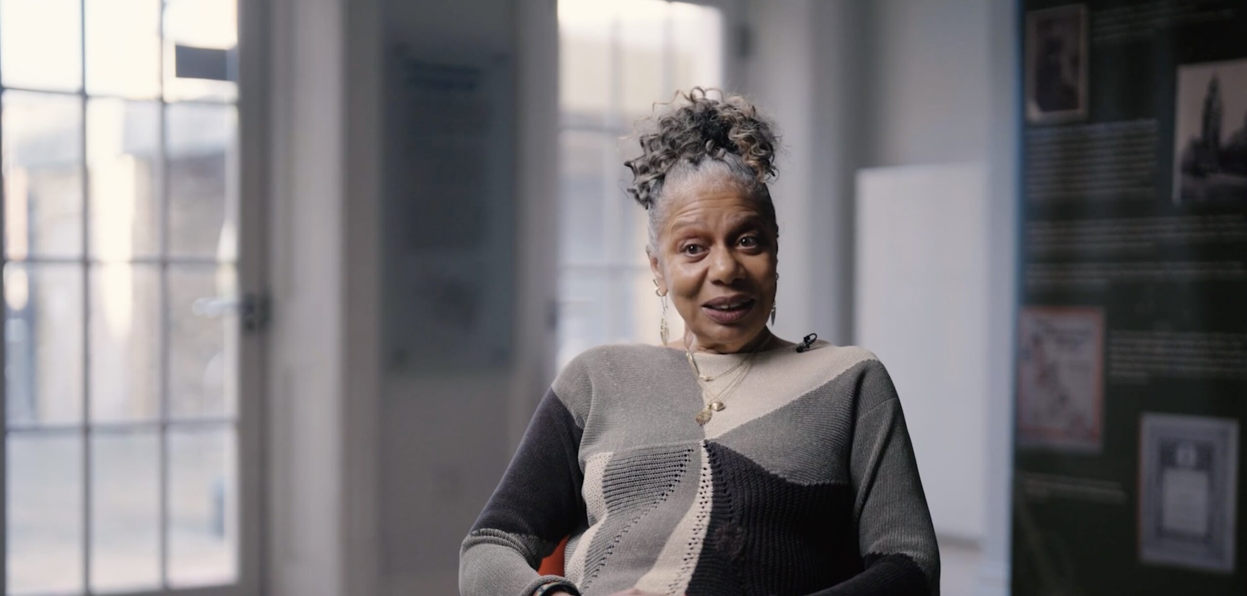 A medium shot of an older Black woman sitting in a chair, appearing to speak while smiling during an interview. She has her hair styled in a high, curly updo with grey streaks. She is wearing a long-sleeved, color-blocked sweater in shades of grey, black, and olive green, layered gold necklaces, and earrings. Her hands are clasped in her lap. The background is a softly blurred indoor setting with a large glass-paned door on the left and an exhibition banner on the right.