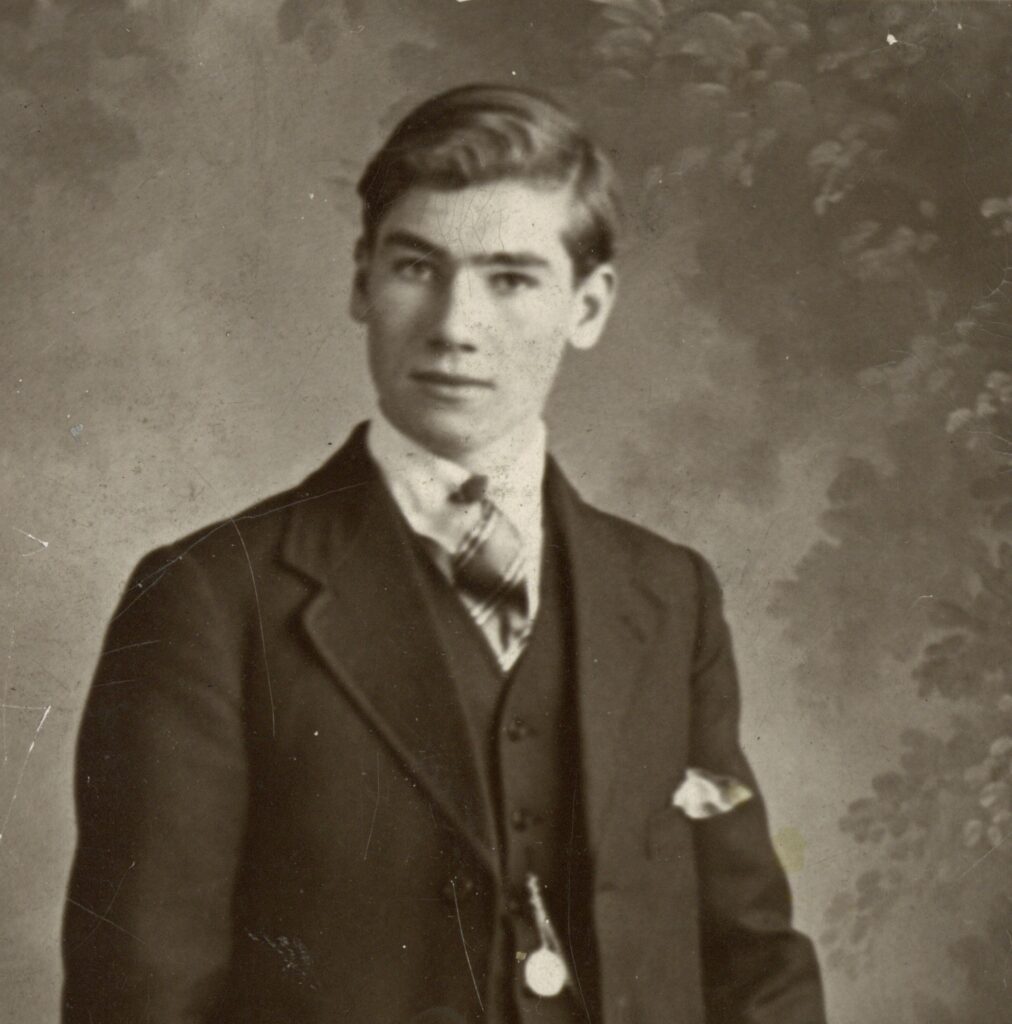 A vintage, sepia-toned studio photograph with a worn, aged border. The portrait shows a young man with combed hair, dressed in a formal dark three-piece suit, white collared shirt, and pattern-knotted tie. A pocket watch chain hangs across his waistcoat, and a pocket square peaks out from his jacket pocket. He stands against a mottled, painted nature backdrop, resting his left hand on the back of a wicker chair.