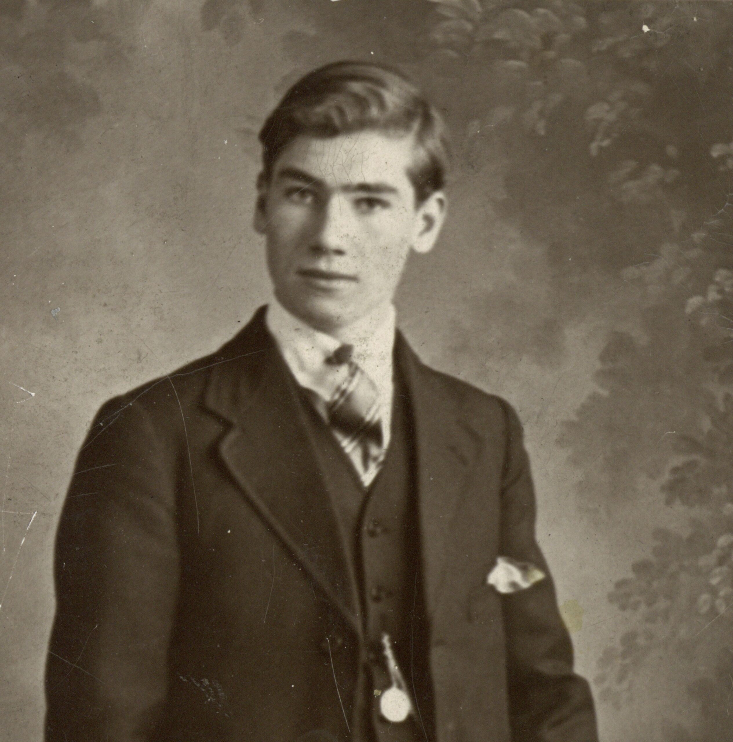 A vintage, sepia-toned studio photograph with a worn, aged border. The portrait shows a young man with combed hair, dressed in a formal dark three-piece suit, white collared shirt, and pattern-knotted tie. A pocket watch chain hangs across his waistcoat, and a pocket square peaks out from his jacket pocket. He stands against a mottled, painted nature backdrop, resting his left hand on the back of a wicker chair.
