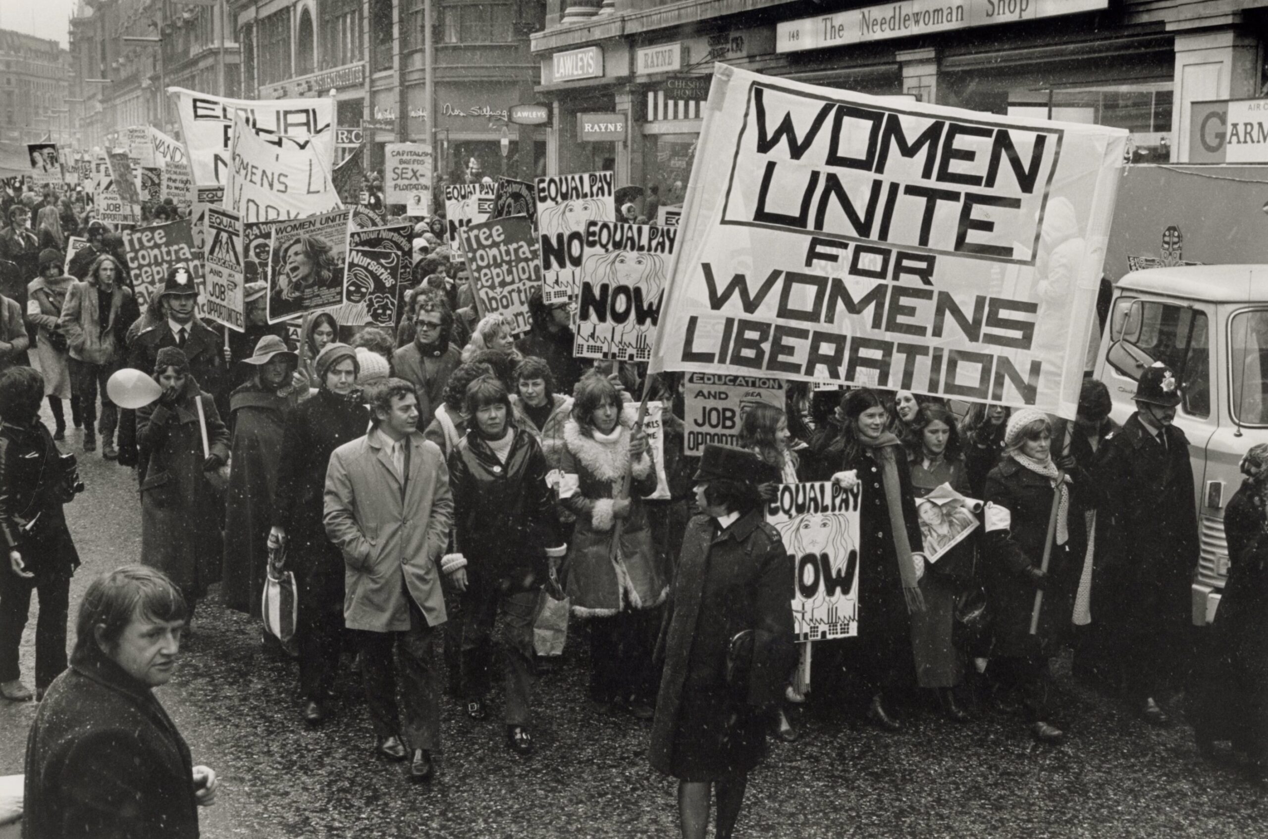 A historic black and white photograph capturing a dense crowd of women marching down a commercial city street during a protest. In the foreground on the right, protestors hold aloft a very large, prominent white banner that reads Women Unite for Womens Liberation in bold black block lettering. Surrounding marchers carry smaller signs advocating for free contraception, equal pay now, and nurseries, while onlookers and traditional storefronts like The Needlewoman Shop line the background street under a light snowfall or rain.