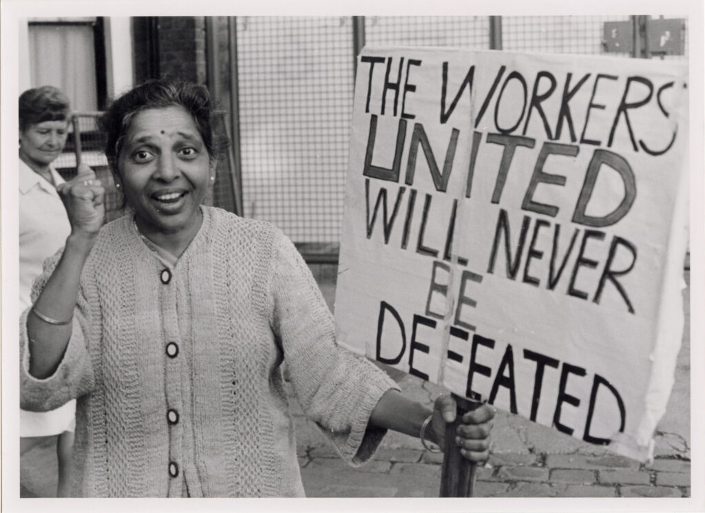 Black and white photograph of an older South Asian woman smiling and raising a clenched fist in solidarity. She holds a large, handwritten protest sign stating 
