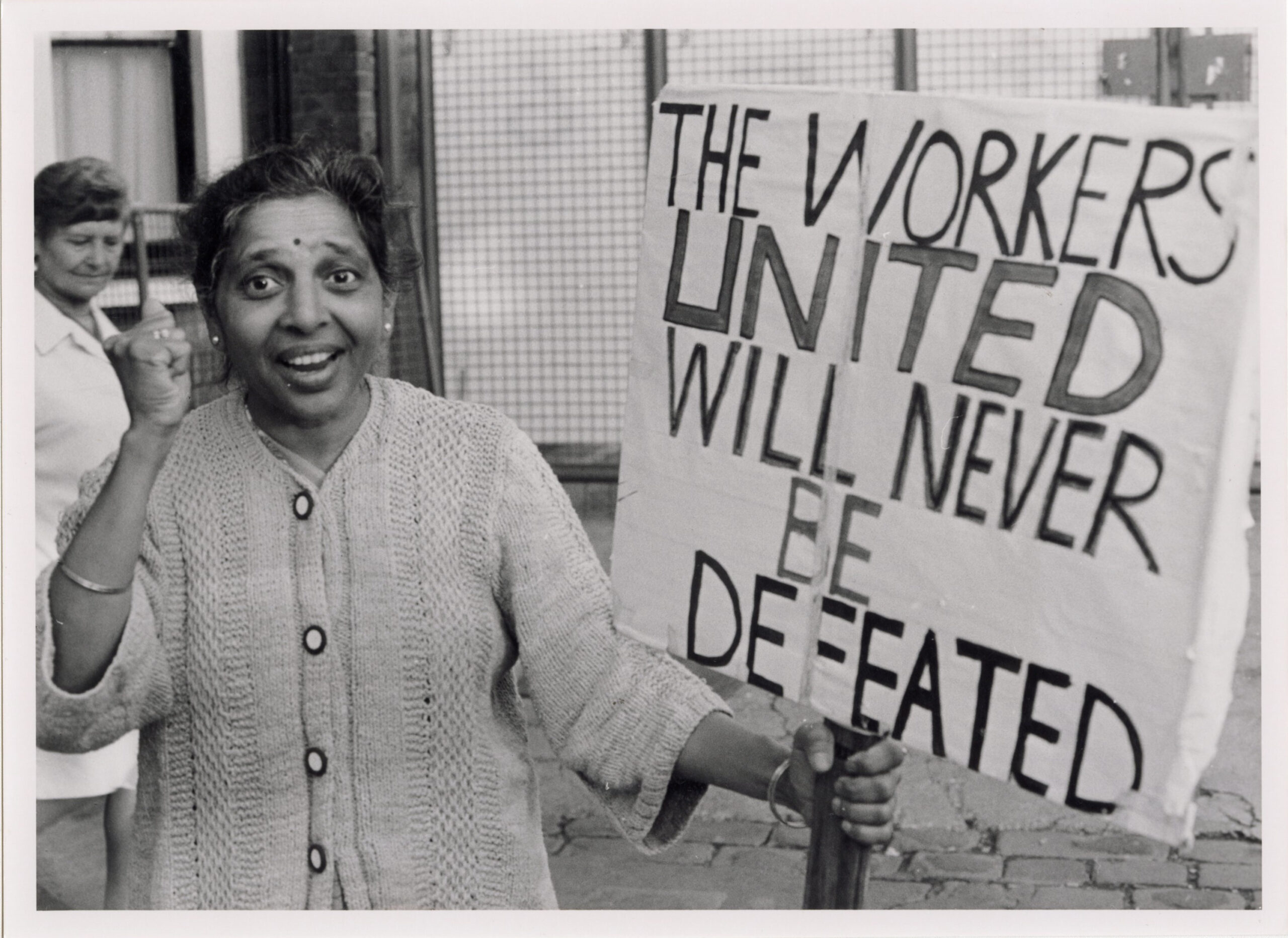 Black and white photograph of an older South Asian woman smiling and raising a clenched fist in solidarity. She holds a large, handwritten protest sign stating 