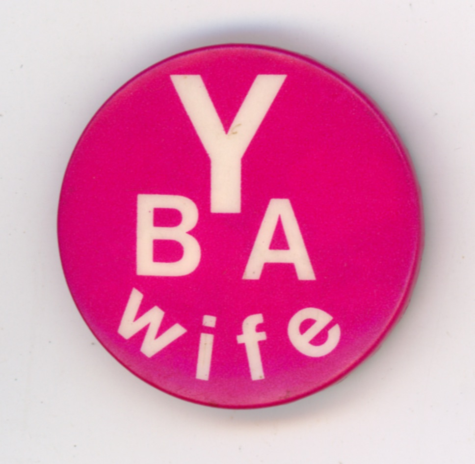 A top-down photograph of a circular pin-back badge with a solid magenta-pink background. Bold, sans-serif white text is arranged vertically in the center to spell out 