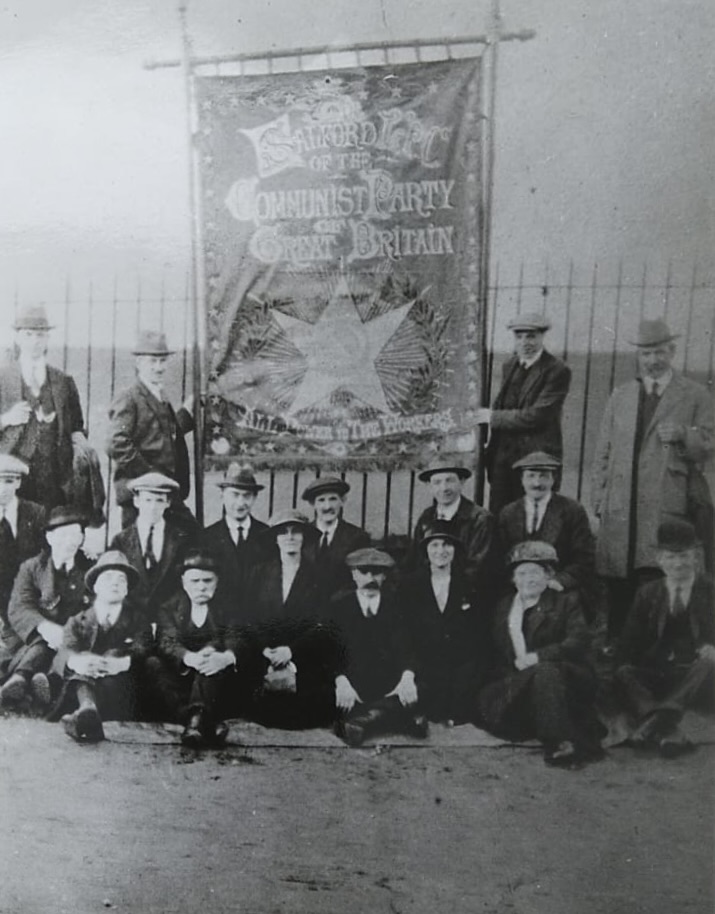 A grainy, black-and-white historical photograph of a group of striking workers or activists posing outside in front of a metal fence. Two men in suits and hats stand holding up a large fabric banner that reads 