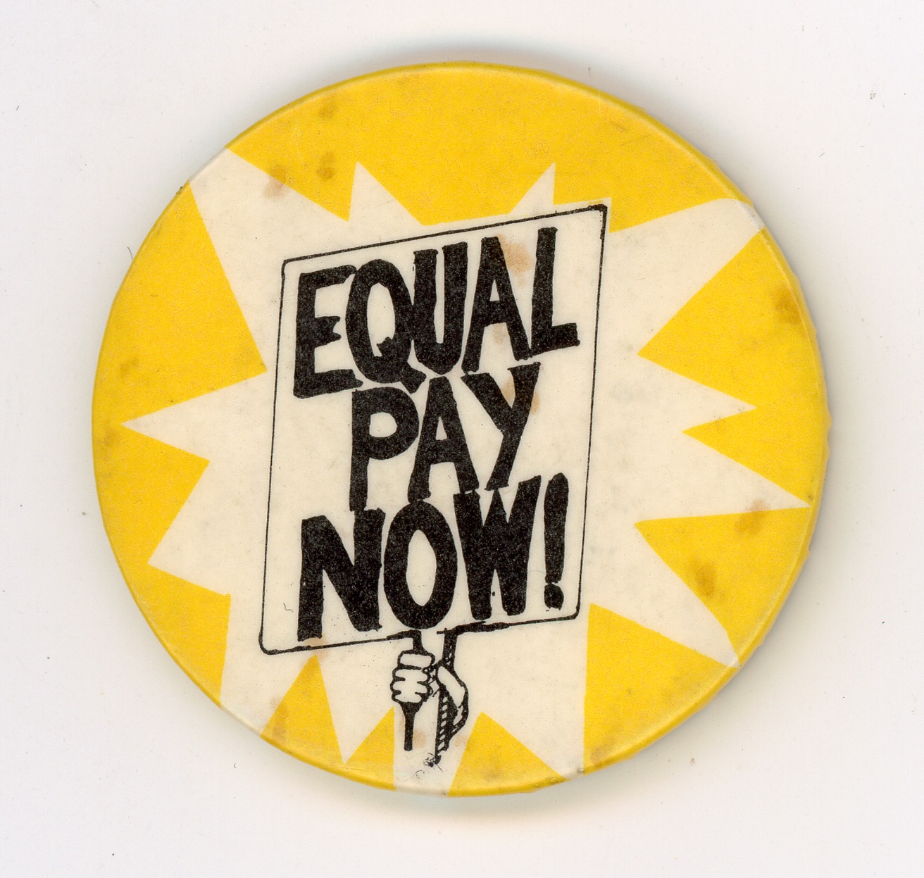 A circular vintage protest badge with a bright yellow background featuring a white starburst design. In the center, a graphic illustration shows a hand holding up a rectangular placard. The placard contains bold, black, hand-drawn text that reads 