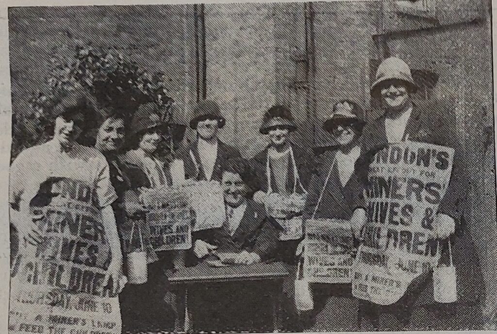 A grainy, archival black and white photograph showing a group of seven women standing together outdoors. They wear hats and coats typical of the 1920s. Two women on the outer edges hold large sandwich-board style signs, while others hold smaller signs and canisters for donations. The visible text on the signs reads 