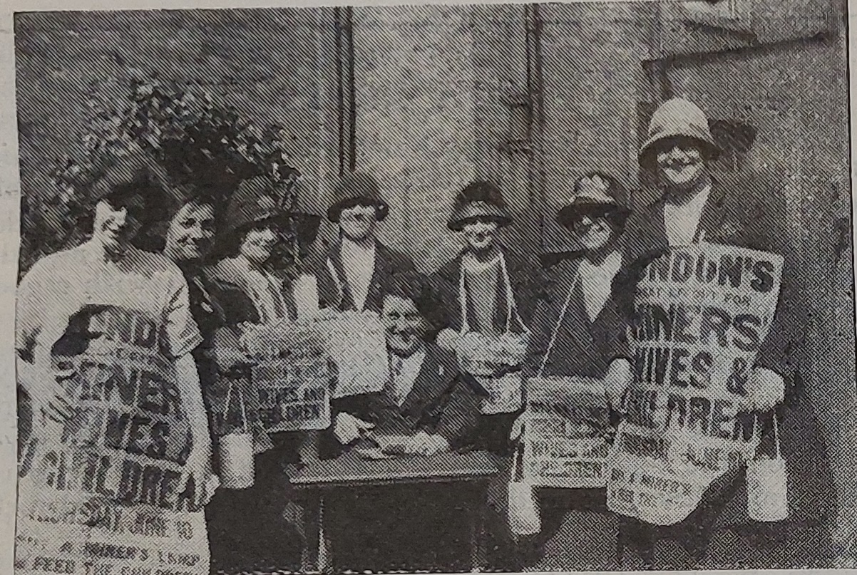 A grainy, archival black and white photograph showing a group of seven women standing together outdoors. They wear hats and coats typical of the 1920s. Two women on the outer edges hold large sandwich-board style signs, while others hold smaller signs and canisters for donations. The visible text on the signs reads 