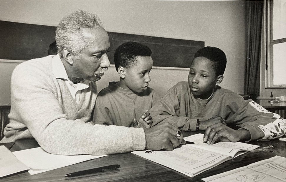 A candid black-and-white classroom photograph. On the left, an older Black man with grey hair, a mustache, and a light-colored V-neck sweater leans forward over a wooden desk. He points with a pen to a page in an open textbook, guiding two young Black schoolboys sitting next to him. The boys, both wearing sweaters, look intently down at the textbook, listening closely. A black pen rests on the table in the foreground, and a blank blackboard stretches across the background wall.