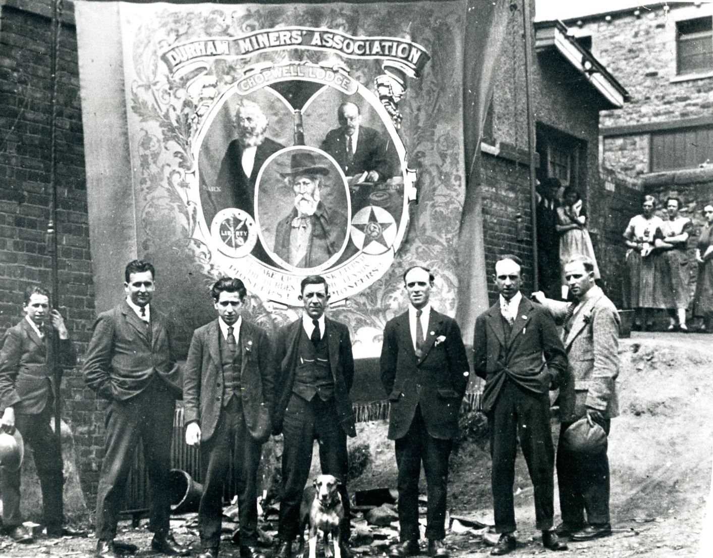 A black-and-white historical photograph of seven working-class men standing outdoors in front of a brick wall, posing before a very large trade union banner. The banner reads 